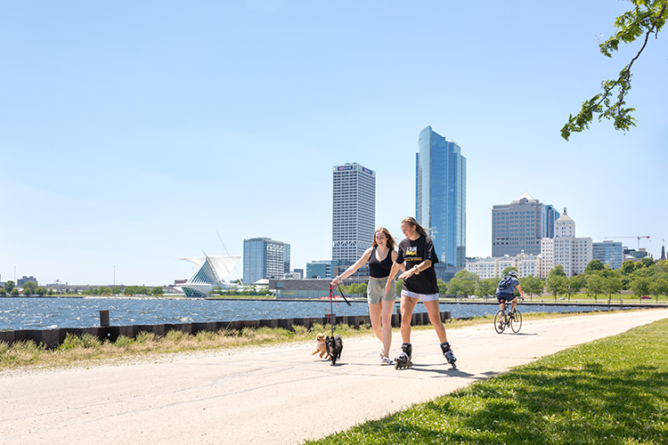 Two UWM female students rollerblading with their dog with the Milwaukee city skyline behind them